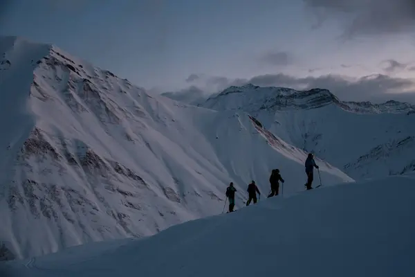 Silhouettes of four skiers climbing a mountain slope against the background of other snowy slopes at dusk