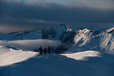 Üç erkek kayakçı, bir dağ tepesinin karlı ovasında kayak yaparken, bir kısmı küçük bir sis şeridiyle kaplanmış bir dağ kitlesinin arka planına karşı çıkıyor.