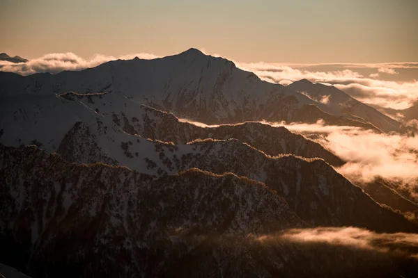 いくつかの山の斜面は 日没に明確な空に対して雪と霧で覆われて互いに平行に配置されました — ストック写真