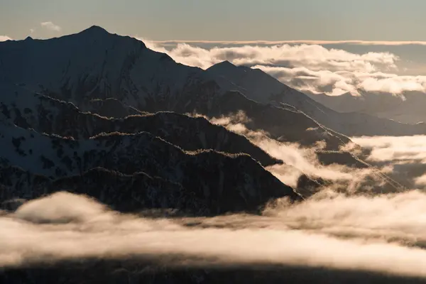 雪で部分的に覆われたシャープな岩の山頂の美しい写真 青空の背景に霧雲が浮かぶ過去 — ストック写真