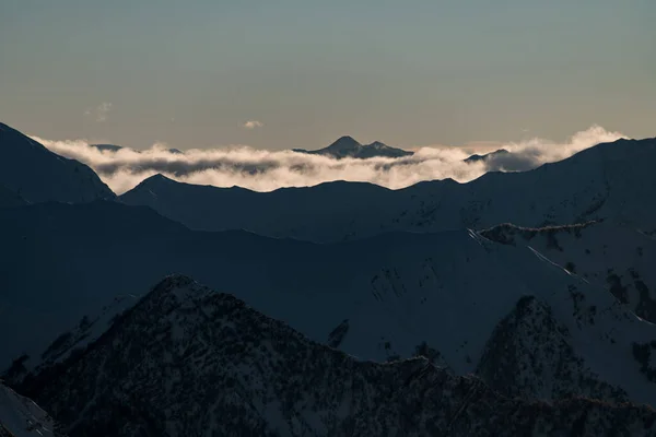 山と合流する明確な青空の背景に対して 霧の流れが遠くに広がる点点のピークを持つダークマウンテンスロープ — ストック写真
