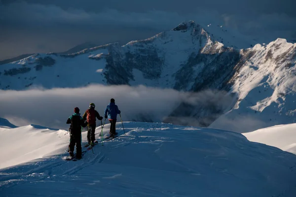 3人の男性スキーヤーが雪に覆われた山頂で次々と歩いていて その背後には霧が山頂の間に広がっている — ストック写真
