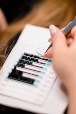 Bir stilistin kadın elleri cımbızla beyaz bir kürsüdeki paletli takma kirpikleri alır.