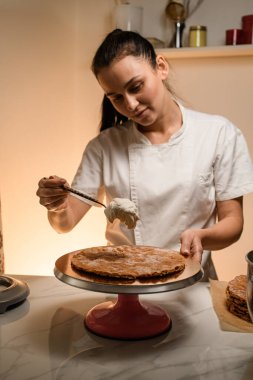 Siyah uzun saçlı, beyaz elbiseli esmer bir kadın pasta yapıyor. Pasta tezgahına krema sürüyorum.