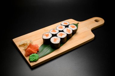 Somon balıklı suşi ruloları, farklı zencefil ve wasabi yeşil yapraklar üzerinde servis edilen siyah bir arka planda.