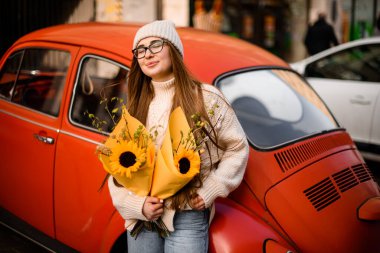 Gözleri kapalı, tatlı bir kadın. Kırmızı bir arabaya yaslanırken elinde iki buket dekoratif ayçiçeği tutuyor.