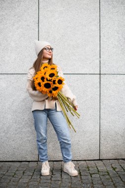 Elinde kocaman bir dekoru olan şapka ve gözlük takan bir kadın kaldırımda dikilip yan tarafa bakarken...