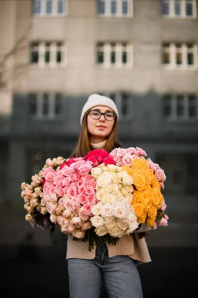 Pretty woman in a hat and glasses holding a huge bouquet of multi-colored roses against the background of a high-rise building