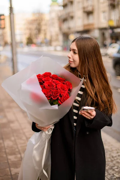 Beautiful brunette woman with large bouquet of red roses for holiday stands on the street. Attractive woman with flowers bouquet . Blurred background