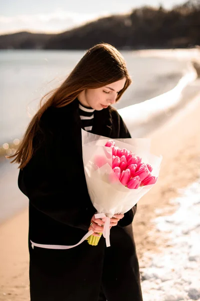 Happy stylish young brunette woman wearing black coat holding bouquet of pink tulip flowers standing by seaside. Elegant caucasian lady in nature, good mood.