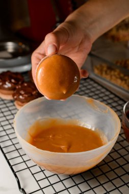 Kadın şekerlemecinin elinde karamel renkli kremaya batırılmış bir badem kurabiyesi var. Metal bir ızgarada duruyor.