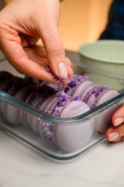 Kadın pasta şefinin ellerini kapayın. Mor badem kurabiyelerini beyaz masaya mor serpiştirilmiş cam kabın içinde krema katmanlarıyla serpin.