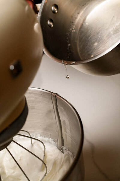 Stand mixer mixes dough for baking macaroon halves on white kitchen table close-up, syrup is added from saucepan