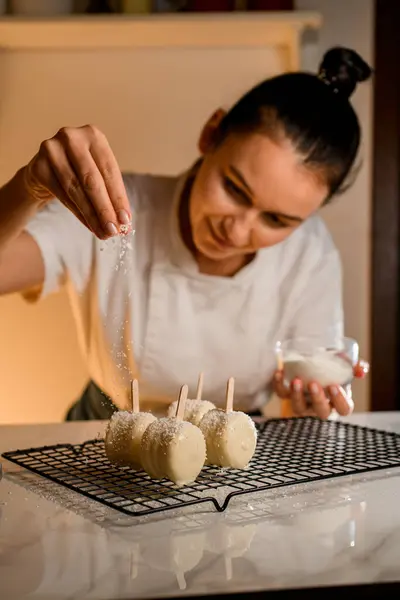 Female confectioner, bending over the table, sprinkles coconut shavings on macaroons on a stick in white cream