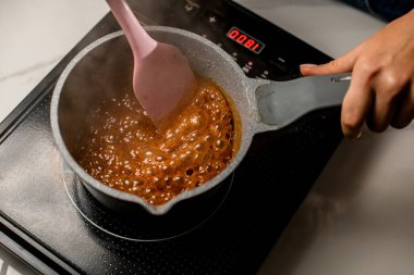 Kadın pastacı, seramik bir tencerede siyah bir indüksiyon sobası üzerinde pembe silikon spatulayla şerbet karıştırıyor.