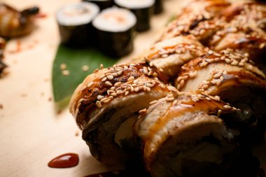 Sosu ve susam tohumlarıyla süslenmiş yılan balığı ile Philadelphia suşi rulolarına odaklan. Maki ve Nigiri bulanık bir arkaplanda görülebilir.