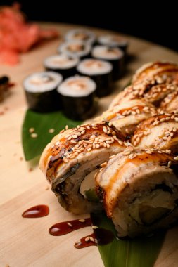 Sosu ve susam tohumlarıyla süslenmiş yılan balığı ile Philadelphia suşi rulolarına odaklan. Maki suşi ve kırmızı zencefil bulanık arka planda ahşap bir yuvarlak tahta üzerinde