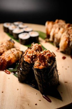 Sos ve susam ile süslenmiş iki silahşöre odaklan. Maki, Nigiri ve Philadelphia suşisi siyah arka planda izole edilmiş ahşap bir tahta üzerinde.