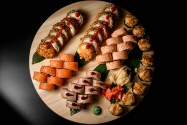 Somon, alabalık ve ton balıklı suşi rulolarının ve siyah arka planda haşlanmış kızarmış ekmeklerin bulunduğu yuvarlak ahşap tahtanın en üst görüntüsü.