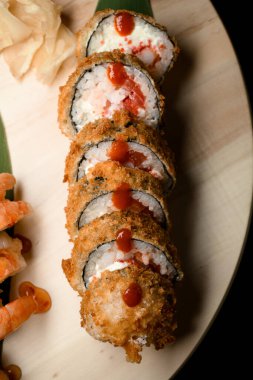 Yengeç dolgulu sıcak suşi rulolarının en üst görüntüsü, beyaz zencefil ve karides rulolarının yanında, açık tahta tahtada siyah masa arkasında.