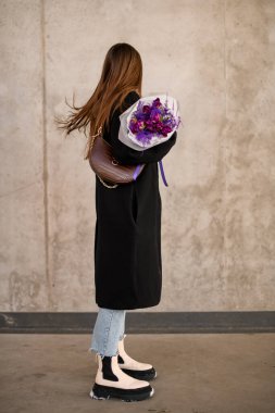 Uzun paltolu, ince bir kadın elinde bir buket mor çiçekle kameradan yüzünü beton bir arka plana çeviriyor.