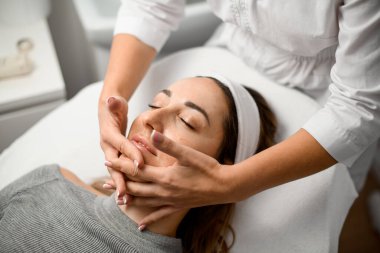 Kozmetik uzmanı, kozmetoloji sandalyesinde uzanan bir müşterinin boynuna elle masaj yapar. Parmaklarını çenesinde çapraz yapar ve kadının arkasında durur.