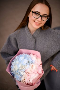 Mutlu gülümseyen genç bir kadın pembe bir hediye paketinde mavi ve pembe ortancalardan oluşan buketi bulanık bir arka planda tutuyor.