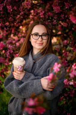 Kartondan bir fincan kahve ve kollarını göğsüne bağlamış bir kadın çiçek açan bir cennet elma ağacının arka planında duruyor.