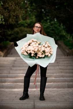 Genç bir kadın merdivenlerde duruyor ve elinde büyük bir buket pembe gülle beyaz bir ambalajda.