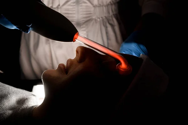 Side view of the face of a woman with a white bandage on her head, to whom a cosmetologist performs a darsonvalization procedure in a beauty salon in low light