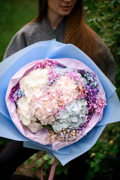 Close-up photo of a huge bouquet of hydrangea with hypophylla in a blue and pink wrapper against a green garden background