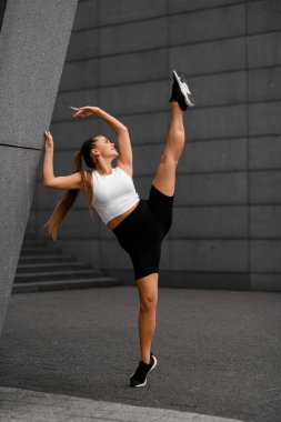 Spor ayakkabılı, şortlu ve tişörtlü bir kız gri kentsel yapılara karşı dışarıda esneme egzersizleri yapıyor..
