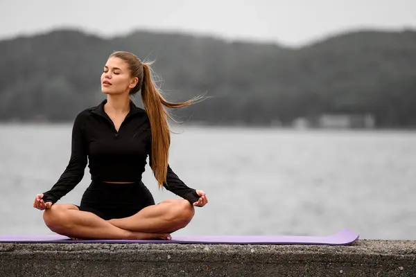 In the serene seashore setting, a young and healthy girl practices her stretching exercises in the lotus position.