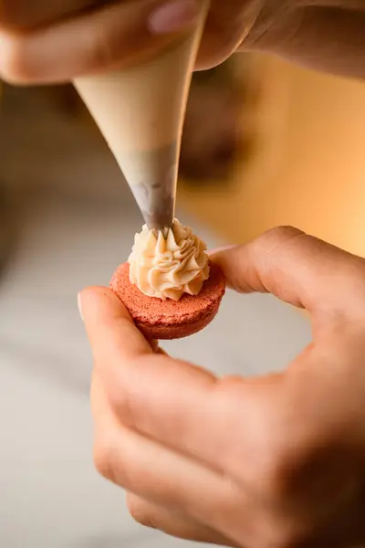 Confectioners hands use a pastry bag to apply white cream to the pink half of the macaroon blank