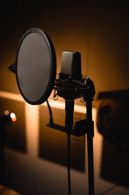 Profesyonel bir mikrofona yakın çekim. Kayıt stüdyosunda kare şeklinde ışıklandırılmış bulanık bir duvar arkaplanı var. Ön plana odaklan.