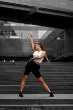 Uzun saçlı, atkuyruklu genç bayan sporcu bacaklarını ayırarak jimnastik yapıyor.