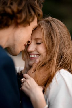 Zarif elbiseli mutlu yeni evliler sıkıca sarılır ve arka plandaki bulanık çekimde alınlarına dokunur.