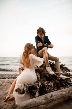 Kadın, beyaz bir elbise içinde, sırtı açık bir şekilde kocasının yanında oturuyor, kocasına bakıyor, kolları bir ağacın köklerinde, göğsünde katlanmış bir şekilde.