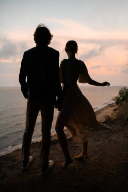 Genç ve narin yeni evliler kumlu deniz kıyısında, denizin ve günbatımının arka planında el ele dururlar.