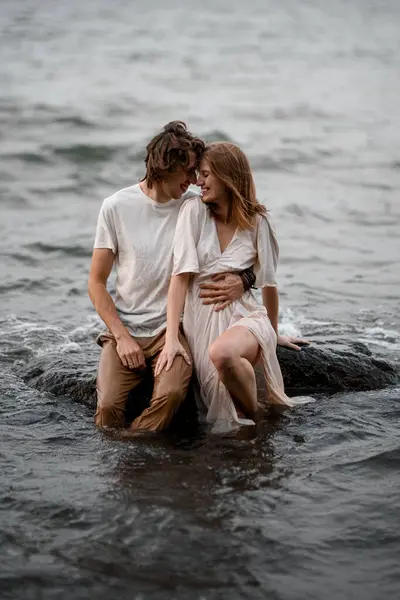 Happy smiling young couple in wet clothes sitting on a rock in the water and hugging each other with their foreheads