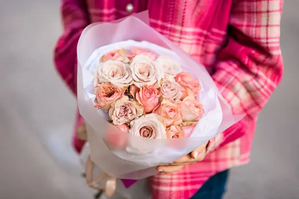 Bouquet of light roses in white matte wrapping paper, in the hands of a young woman in a pink shirt on a blurred background