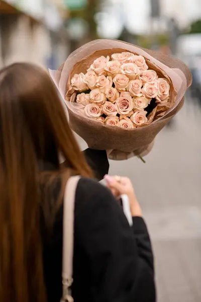 彼女の前に高いクラフト紙に包まれた桃のバラの大きな花束を保持している若い女性 選択的な焦点 バックビュー ストック画像