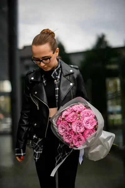 黄色いラッピングペーパーに包まれたピンクのバラの花束を身に着けている若い美しい女性は ぼやけた街並みの背景を見下ろす ストックフォト