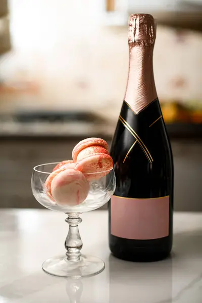 ピンクのマカロンの高いガラスは台所のテーブルの上に立っています シャンパンのボトルはぼやけた背景のそれの隣に立っています ストック画像