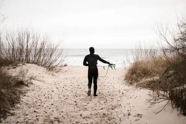 若い男性サーファー ウェットスーツを着て 茂みの間の砂の上を歩き 彼のサーフボードを手に持って 海と明るい空の背景に対して ストック写真
