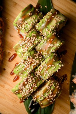 Top view of sushi wrapped in green decorated with white sesame and thick light brown sauce on a long green leaf on a wooden board clipart