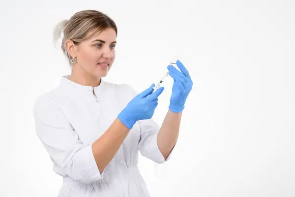 白いオーバーオールと青いゴム製手袋の美容分野の女性化粧品学者は サロンプロシージャのための注射器に化粧品を描きます ストック画像