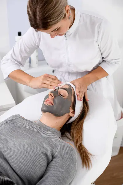 白いオーバーオールの美容分野のスペシャリストは 灰色のセーターで軽い髪を持つ美しい女性のための顔のマスクを作ります ストック画像
