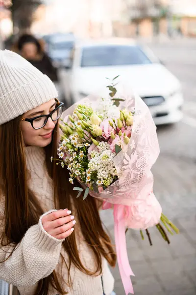 大きな花束を持つ街の通りに立っている黒いフレームのメガネでまっすぐな髪とフェアな肌の少女 ストック写真