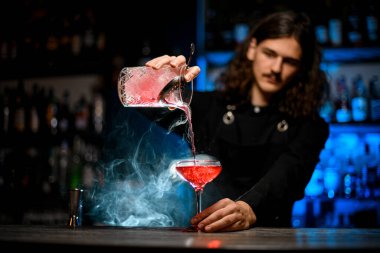 Uzun kıvırcık saçlı barmen kokteyl bardağına soğuk kırmızı kokteyl doldurmayı bitirdi.